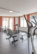 Salle de fitness baignée de lumière à l'hôtel Alpenblick avec appareils de cardio et de musculation modernes, grandes fenêtres et serviettes jaunes offertes.